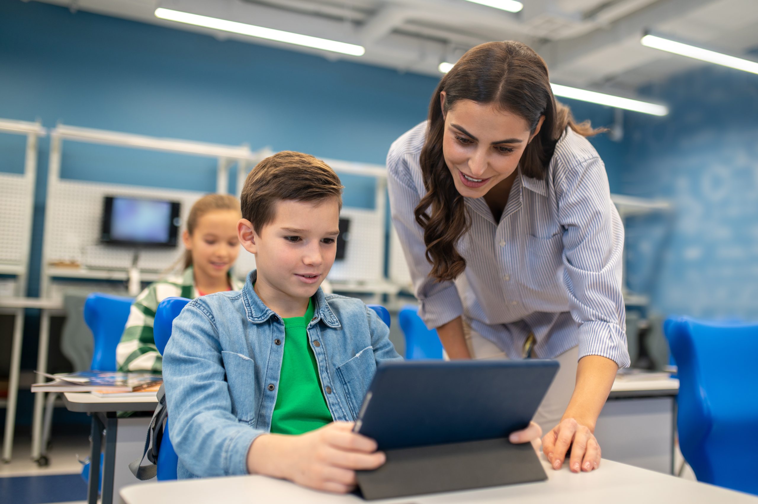 Education Queue management software Interesting here. Interested smiling young woman looking into tablet of student boy sitting at desk in school illuminated classroom