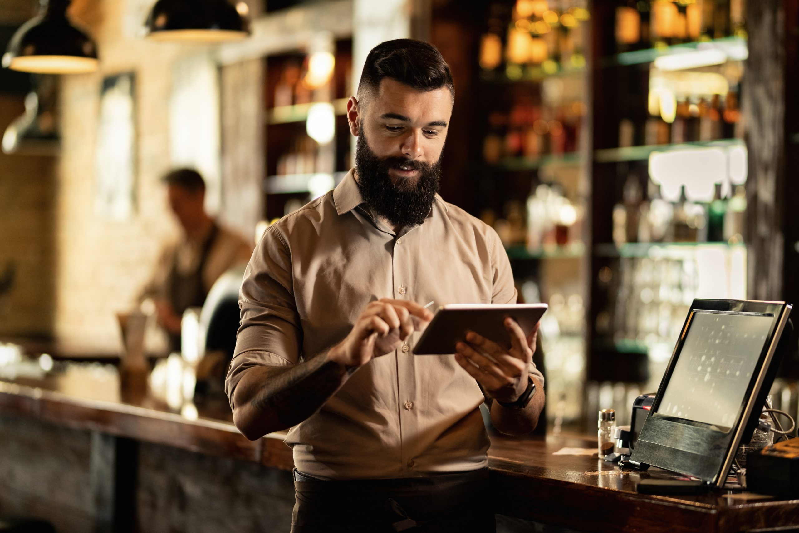 Smiling barista using digital tablet while working in a bar. Young smiling waiter using touchpad while working in a pub.